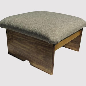 May include: A small, rectangular footstool with a gray fabric cushion and a wooden frame. The frame is made of dark brown wood with a natural grain. The footstool is designed with a simple, minimalist aesthetic.