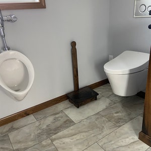 May include: A white urinal with a chrome faucet and a wooden step stool in a bathroom with gray tile flooring.