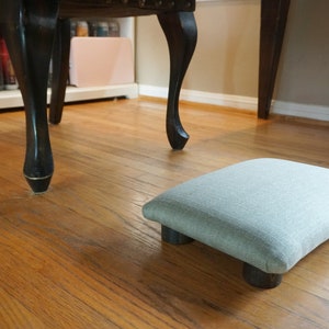 Puede incluir: Un reposapiés de tela verde claro con cuatro patas de madera. El reposapiés está sobre un suelo de madera.