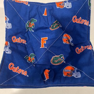May include: Blue fabric pot holder with a white stitched border. The fabric has a repeating pattern of orange and green alligators, football helmets, and the word "Gators". The center of the pot holder has a large white "F" in the center.