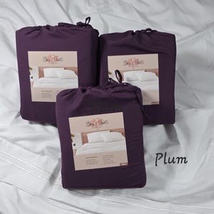 Puede incluir: Tres juegos de sábanas color ciruela, cada uno en una bolsa con cordón, se exhiben sobre una cama blanca. Cada bolsa tiene una etiqueta con el texto "Stacy's Sheets" y una imagen de una cama. La palabra "Plum" está escrita en cursiva.