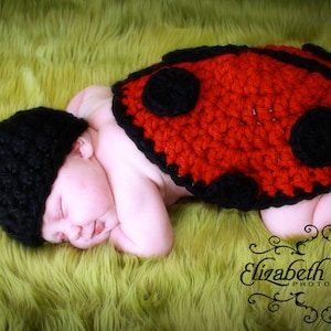 May include: A sleeping baby wearing a red and black ladybug costume. The costume is crocheted and has black spots. The baby is lying on a green fuzzy surface.