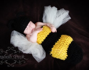 May include: A baby wearing a crocheted yellow and black bee costume with a white tulle skirt. The baby is sleeping and has a black crocheted bee hat on.