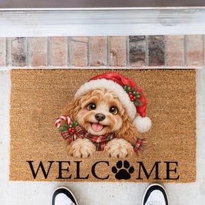 May include: A rectangular tan coir doormat featuring a graphic of a curly-haired puppy wearing a red Santa hat and a plaid scarf while holding a candy cane. The text WELC(paw print)ME is printed in black capital letters at the bottom of the mat.