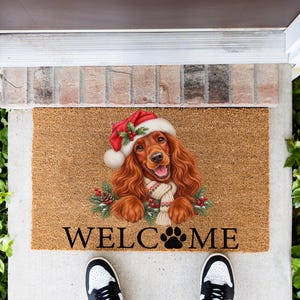 May include: A rectangular tan coir doormat featuring a brown spaniel dog wearing a red and white Santa hat and a white knit scarf. The dog is surrounded by green pine branches and red berries. The text WELC(paw print)ME is printed in black.