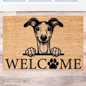 May include: A rectangular doormat with a dog design. The mat is tan with a black illustration of a dog's head and paws, and the word "WELCOME" in large black letters. A paw print replaces the "O" in "WELCOME".