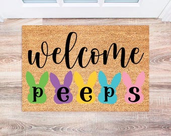 May include: A brown coir doormat with the text "welcome" in black cursive lettering. Below the text are five colorful bunny-shaped figures with the letters "p e e p s" in black.