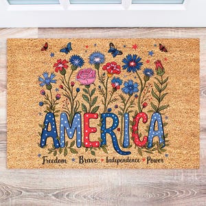 May include: A brown coir doormat with the word "AMERICA" in colorful letters with white polka dots. The letters are surrounded by a floral design with red, white, and blue flowers. The text "Freedom - Brave - Independence - Power" is printed below the word "AMERICA".