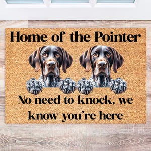 May include: A brown and white doormat with the text "Home of the Pointer. No need to knock, we know you're here." Two brown and white pointer dogs are pictured on the mat.