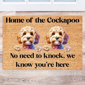 May include: A brown doormat with the text "Home of the Cockapoo. No need to knock, we know you're here." Two brown and white Cockapoo dogs are peeking out from behind the text.