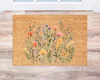 May include: A brown coir doormat with a floral design featuring orange, yellow, and blue flowers. The flowers are arranged in a cluster and are surrounded by green leaves.