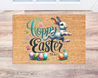 May include: A rectangular doormat with a light brown, textured surface. The mat features a white rabbit leaping above the words "Hoppy Easter to All" in teal and black script. Colorful Easter eggs and paw prints are also depicted.