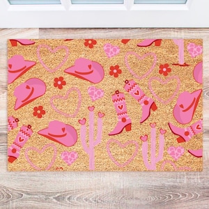 May include: A pink and red doormat with a cowboy boot, cactus, and heart pattern. The doormat is made of coir and has a natural brown color.