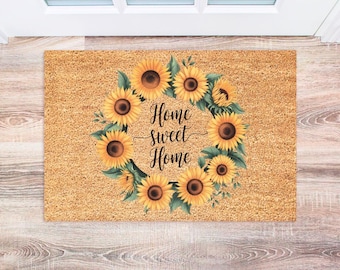 May include: A brown coir doormat with a wreath of yellow sunflowers and green leaves. The text "Home Sweet Home" is written in black in the center of the wreath.