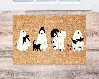 Könnte beinhalten: Eine braune Kokosfaser-Fußmatte mit Halloween-Motiv, auf der fünf weiße Geister mit schwarzen Katzen zu sehen sind. Die Geister halten die Katzen in ihren Armen. In der Mitte der Fußmatte befindet sich ein Kürbis mit Süßigkeiten.