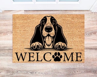 May include: A rectangular doormat with a light brown background and a black and white illustration of a Basset Hound. The word "WELCOME" is printed in black, with a paw print replacing the "O".