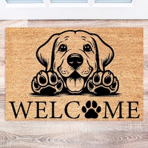 May include: A rectangular doormat with a smiling dog graphic and the word "WELCOME" in black. The dog's paws are also featured. The mat is made of a natural fiber material.