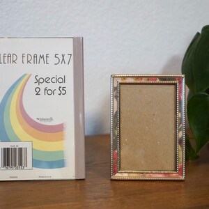 May include: A clear 5x7 inch photo frame next to a decorative metal frame with a floral design. The clear frame is in its original packaging, which has a rainbow graphic and the text "CLEAR FRAME 5X7".