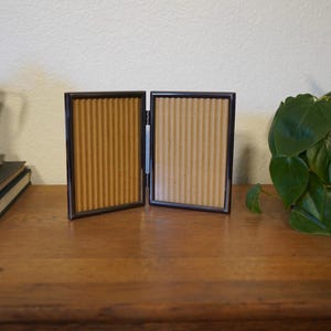 May include: A hinged, two-panel picture frame with a dark frame and a corrugated cardboard backing. The frame is open, displaying the textured backing. The frame sits on a wooden surface, with a plant in the background.