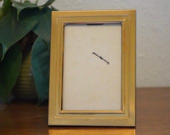 May include: A rectangular gold-coloured picture frame with a cream-coloured interior. The frame has a simple, elegant design and is sitting on a wooden surface. A green plant is visible in the background.