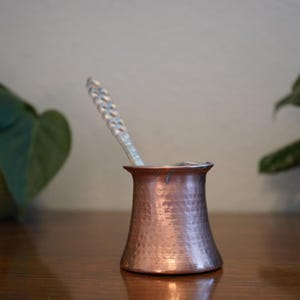 May include: A copper cezve, a traditional Turkish coffee pot, with a hammered texture. A silver spoon rests inside the pot. The cezve sits on a wooden surface, with green plants in the background.