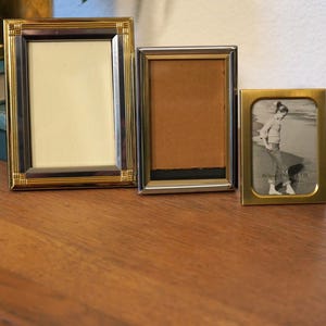 May include: Three picture frames of varying sizes sit on a wooden surface. The largest frame has a gold and brown border, the middle frame has a silver border, and the smallest frame is gold. The smallest frame contains a black and white photo.