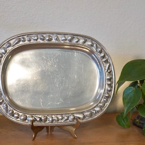 May include: Oval silver-toned serving tray with a decorative border of raised, rounded shapes. The tray sits on a small, gold-toned stand. A green plant is visible in the background.