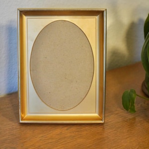 May include: A rectangular gold-toned picture frame with an oval opening. The frame has a thin, metallic border and a cream-colored interior. The oval space is filled with a light brown backing.