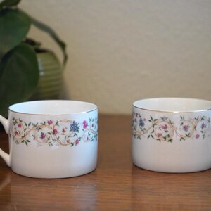 May include: Two white teacups with floral and gold trim. The cups have a curved handle and a decorative band of pink, blue, and green flowers and vines. The cups are on a wooden surface.