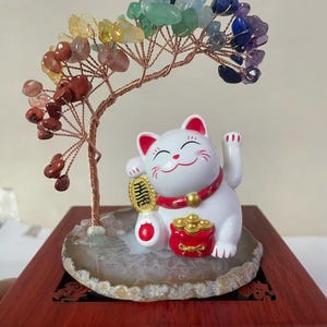 May include: A white Maneki Neko cat figurine with a red collar and gold accents, holding a gold coin and a red pot. A wire tree with colorful gemstone leaves sits above the cat on a wooden base, creating a unique decorative piece.