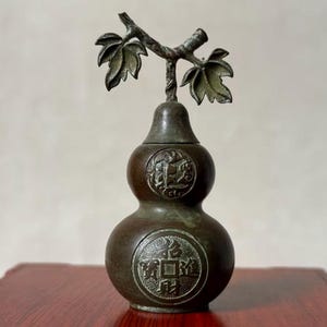 May include: A bronze-toned metal gourd-shaped vessel with a removable lid topped by a stem with two leaves. The body features two circular embossed medallions, with the lower one displaying four Chinese characters and the upper one a floral motif.
