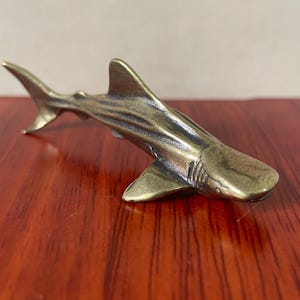 May include: A bronze-colored shark figurine with detailed fins and body ridges. The shark is positioned on a reddish-brown wooden surface, showcasing its metallic sheen. The figurine is a decorative item, possibly a collectible or a nautical-themed piece.