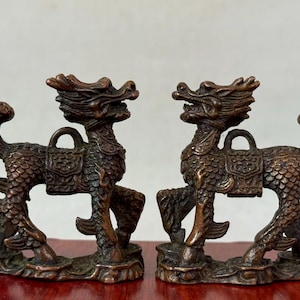 May include: A pair of bronze-coloured mythical creature figurines, each featuring a dragon-like head, a scaled body, a saddle with a loop on the back, and a curved tail. The figures stand on textured bases with intricate detailing throughout.