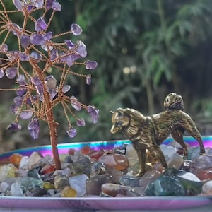 May include: A decorative arrangement featuring a copper wire tree adorned with purple amethyst crystals. A small bronze dog figurine stands on a bed of colorful stones within an iridescent dish. The background is blurred.