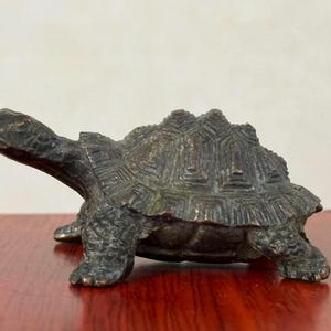 May include: Bronze turtle figurine with detailed shell and textured skin. The sculpture is a dark, antique bronze color and sits on a wooden surface. The turtle's head is extended, and its legs are visible.