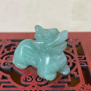May include: A carved jade figurine of a mythical creature in a seated position. The figurine is a light green color and has detailed features. The creature has a smiling expression and is resting on a red wooden surface.