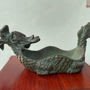 May include: A bronze-toned metal dish shaped like a dragon with a detailed head, scales along the body, and an upturned tail. The vessel features a weathered, dark green patina finish and an open, curved center for holding small items.