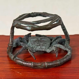 May include: A dark bronze-toned metal crab figurine featuring a circular frame constructed from segmented bamboo-like stalks. The crab is positioned in the centre of the circular base, with its legs and claws extending outward toward the frame.