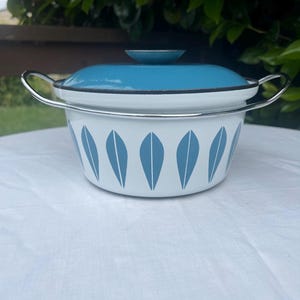 May include: A vintage enamelware pot with a lid. The pot is white with a pattern of blue leaf shapes. The lid and handle are blue and silver. The pot is sitting on a white surface.