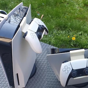 May include: A white PlayStation 5 console with a white controller attached to a stand. The console is on a gray surface.