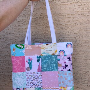 May include: A patchwork tote bag with a variety of colorful patterns, including rainbows, cacti, seashells, and pineapples. The bag has white straps and the text "Handmade with love" is visible on the front.