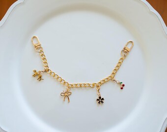 May include: A gold chain with a variety of charms, including a bow, a flower, a cherry, and a small animal figure. The chain has two clasps on either end.