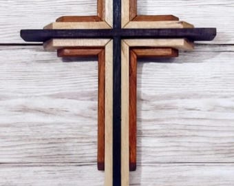 May include: A layered wooden cross featuring a central vertical piece of dark black wood, flanked by lighter tan and medium brown wooden segments arranged in a geometric, multi-dimensional structure with clean, straight edges and a smooth finish.