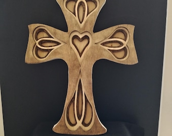 May include: Wooden cross with a heart and floral design. The cross is made of light-colored wood with darker wood accents. The design includes a heart in the center and floral shapes on the arms of the cross. The cross is displayed against a dark background.