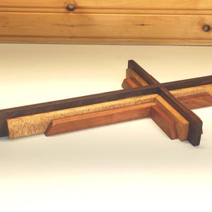 May include: A layered wooden cross with varying shades of brown and tan. The cross is constructed from several pieces of wood, creating a dimensional effect. The design features a dark brown outer layer, with lighter wood tones in the center.