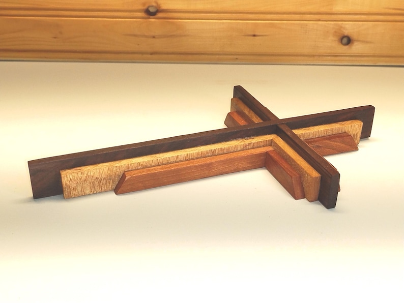 May include: A layered wooden cross with varying shades of brown and tan. The cross is constructed from several pieces of wood, creating a dimensional effect. The design features a dark brown outer layer, with lighter wood tones in the center.