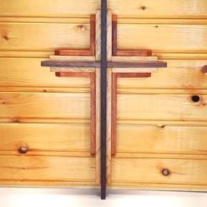 May include: A wooden cross with layered design in shades of brown, red, and natural wood tones. The cross is mounted on a light brown wooden plank background. The cross is a religious symbol.