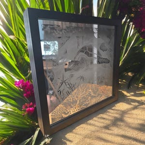Puede incluir: Una obra de arte enmarcada que presenta un diseño detallado e intrincado de una tortuga marina y un arrecife de coral. La obra de arte está en un marco negro y se muestra sobre un fondo de follaje verde.