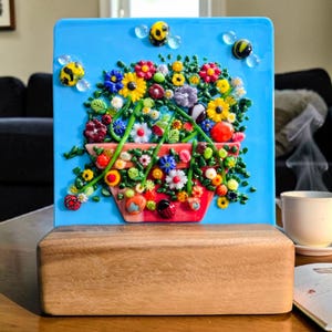 May include: A rectangular fused glass art piece featuring a vibrant floral arrangement in a red pot against a bright blue background, accented with small glass bees and clear droplets, all mounted on a solid rectangular wooden base stand.