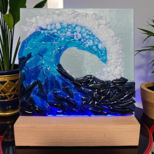 May include: A square glass art piece depicting a breaking ocean wave. The wave is crafted from shades of blue and white glass, with a dark blue base. It sits on a rectangular wooden base with a blue LED light. The artwork is a decorative home accent.